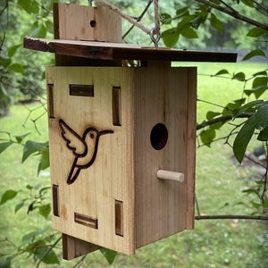 May include: A wooden birdhouse with a hummingbird design, hanging from a tree branch. The birdhouse has a rectangular shape with a small perch and a circular entrance. The hummingbird is etched into the wood.