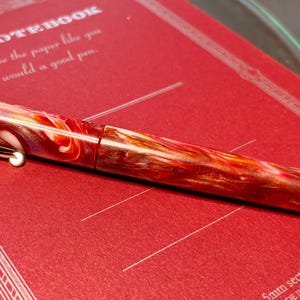 Puede incluir: Una vibrante pluma estilogr&aacute;fica con un patr&oacute;n arremolinado de rojo, naranja y blanco descansa sobre un cuaderno rojo. La pluma tiene un clip y detalles dorados. El cuaderno tiene el texto "C.D. NOTEBOOK" y "Choose the paper like you would a good pen."