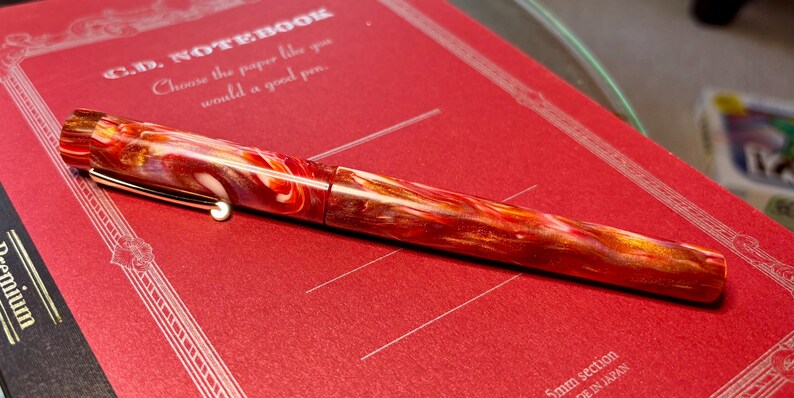 Puede incluir: Una vibrante pluma estilogr&aacute;fica con un patr&oacute;n arremolinado de rojo, naranja y blanco descansa sobre un cuaderno rojo. La pluma tiene un clip y detalles dorados. El cuaderno tiene el texto "C.D. NOTEBOOK" y "Choose the paper like you would a good pen."