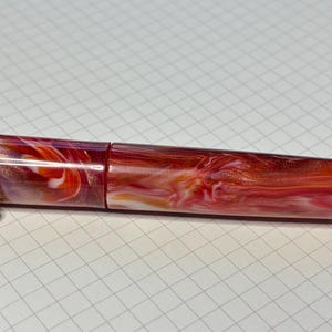 Puede incluir: Pluma estilogr&aacute;fica con dise&ntilde;o veteado en tonos rojos, naranjas y blancos. El bol&iacute;grafo tiene un clip dorado y un acabado pulido. Descansa sobre papel cuadriculado blanco. El bol&iacute;grafo mide unos 14 cm de largo.