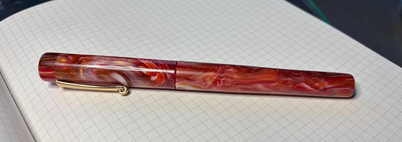 Puede incluir: Pluma estilogr&aacute;fica con dise&ntilde;o veteado en tonos rojos, naranjas y blancos. El bol&iacute;grafo tiene un clip dorado y un acabado pulido. Descansa sobre papel cuadriculado blanco. El bol&iacute;grafo mide unos 14 cm de largo.