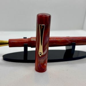 Puede incluir: Una pluma estilogr&aacute;fica veteada en rojo y blanco con plum&iacute;n dorado y clip dorado. La pluma se exhibe sobre un soporte negro. La tapa de la pluma est&aacute; separada y colocada al lado.