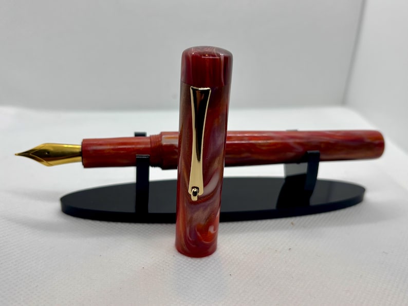 Puede incluir: Una pluma estilogr&aacute;fica veteada en rojo y blanco con plum&iacute;n dorado y clip dorado. La pluma se exhibe sobre un soporte negro. La tapa de la pluma est&aacute; separada y colocada al lado.