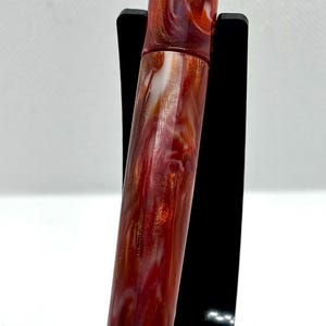 Puede incluir: Pluma estilogr&aacute;fica artesanal con un cuerpo de resina veteada roja, blanca y dorada. La pluma presenta adornos dorados y un clip dorado. La pluma se exhibe sobre un soporte negro sobre un fondo blanco. Un instrumento de escritura.