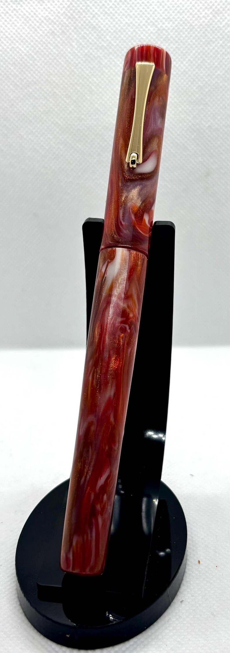 Puede incluir: Pluma estilogr&aacute;fica artesanal con un cuerpo de resina veteada roja, blanca y dorada. La pluma presenta adornos dorados y un clip dorado. La pluma se exhibe sobre un soporte negro sobre un fondo blanco. Un instrumento de escritura.