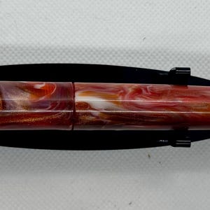 Puede incluir: Una pluma estilogr&aacute;fica de resina veteada con un clip dorado. El cuerpo de la pluma presenta una mezcla de tonos rojos, blancos y marrones. La pluma se exhibe sobre un soporte negro.