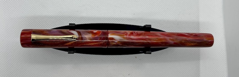 Puede incluir: Una pluma estilogr&aacute;fica de resina veteada con un clip dorado. El cuerpo de la pluma presenta una mezcla de tonos rojos, blancos y marrones. La pluma se exhibe sobre un soporte negro.