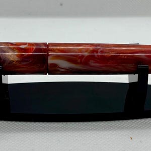 Puede incluir: Una pluma estilogr&aacute;fica veteada en rojo y blanco con detalles dorados, expuesta sobre un soporte ovalado negro. El cuerpo de la pluma presenta un patr&oacute;n arremolinado de rojo, blanco y toques de naranja. Es un instrumento de escritura.