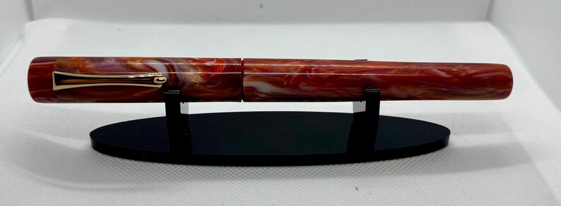 Puede incluir: Una pluma estilogr&aacute;fica veteada en rojo y blanco con detalles dorados, expuesta sobre un soporte ovalado negro. El cuerpo de la pluma presenta un patr&oacute;n arremolinado de rojo, blanco y toques de naranja. Es un instrumento de escritura.