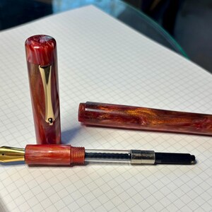 Puede incluir: Pluma estilogr&aacute;fica desmontada con cuerpo y capuch&oacute;n veteados en rojo y marr&oacute;n. Se ve el plum&iacute;n dorado, junto con un clip dorado en el capuch&oacute;n. La pluma descansa sobre papel cuadriculado blanco.