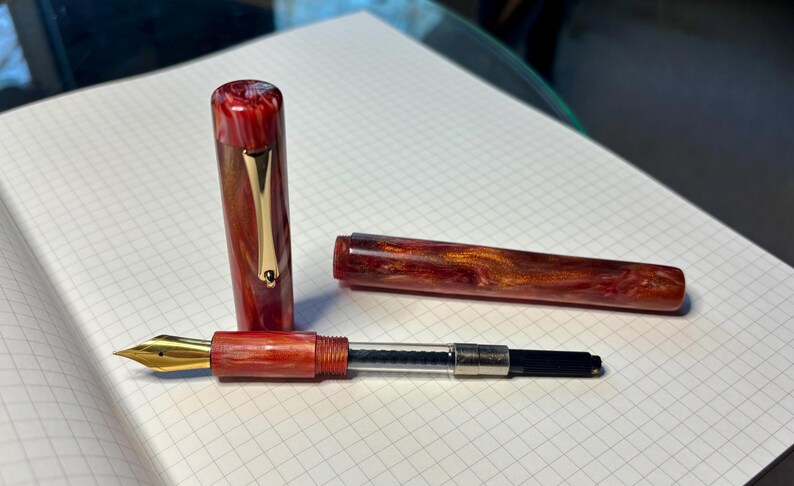 Puede incluir: Pluma estilogr&aacute;fica desmontada con cuerpo y capuch&oacute;n veteados en rojo y marr&oacute;n. Se ve el plum&iacute;n dorado, junto con un clip dorado en el capuch&oacute;n. La pluma descansa sobre papel cuadriculado blanco.