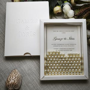May include: A white wedding invitation set featuring a framed invitation filled with pearls. The invitation includes the names "Gamze & Idris" and event details. The invitation is in a white box with embossed text.