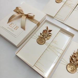 May include: Ivory wedding invitation set in a box with a gold ribbon and bow. The invitation features gold foil details, including initials and the date 22.09.2024. A gold leaf and seal embellish the invitation.