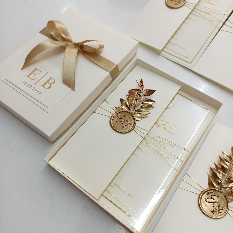 May include: Ivory wedding invitation set in a box with a gold ribbon and bow. The invitation features gold foil details, including initials and the date 22.09.2024. A gold leaf and seal embellish the invitation.