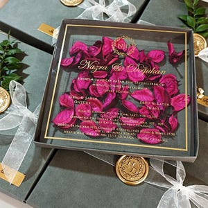 May include: A clear acrylic wedding invitation with pink flower petals inside, featuring gold text and a gold seal. The invitation is in a dark gray box tied with a white ribbon. The text includes names and event details.