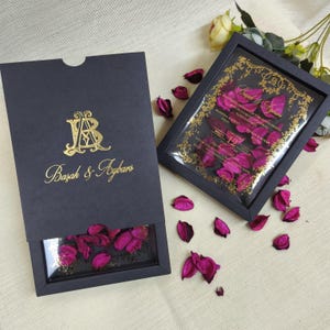 May include: Two black wedding invitation boxes with gold lettering and a clear window displaying pink flower petals. One box is open, revealing the petals. The other box is a framed invitation with gold decorative accents and text.