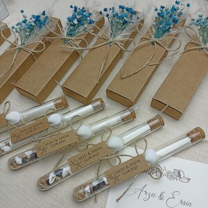 Peut inclure: Des cadeaux de mariage avec de petits tubes en verre bouchés de liège, remplis de sable et de coquillages, attachés avec de la ficelle. Chaque tube porte une étiquette "ARZU & ERSIN 12.08.2023". Des boîtes marron avec des fleurs bleues sont également incluses.