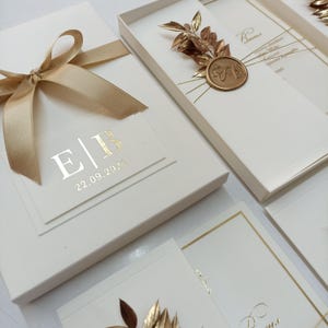 May include: Ivory-colored invitation boxes with gold accents. One box is tied with a gold ribbon. The boxes feature gold foil lettering with the initials "E|B" and the date "22.09.2024". Some boxes have gold leaf and a gold wax seal.
