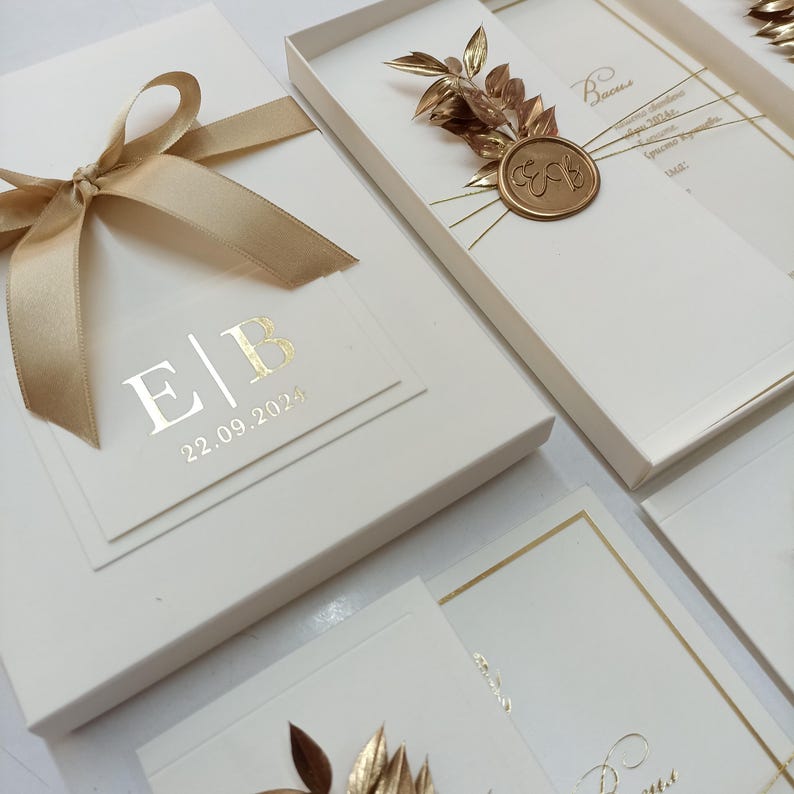 May include: Ivory-colored invitation boxes with gold accents. One box is tied with a gold ribbon. The boxes feature gold foil lettering with the initials "E|B" and the date "22.09.2024". Some boxes have gold leaf and a gold wax seal.