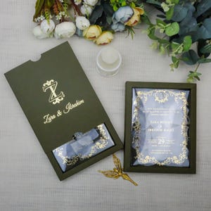 May include: An olive green wedding invitation suite with gold foil details. The invitation box is open, revealing a clear acrylic invitation with gold floral accents. The names "Zara & Ibrahim" are displayed in gold script.