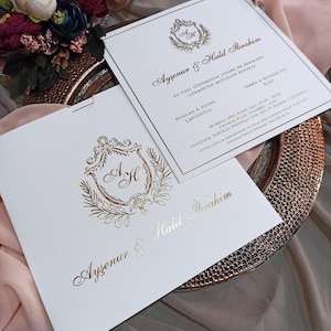 Può includere: Eleganti inviti di nozze bianchi con scritte e dettagli dorati. Gli inviti presentano un stemma decorativo con le iniziali "A H" e sono presentati su un piatto decorativo dorato. Il tema generale è formale e celebrativo.