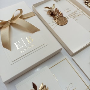 May include: Elegant ivory wedding invitations with gold foil accents. The invitations feature a gold ribbon, initials, and the date "22.09.2024". Some invitations are in boxes, adorned with gold leaf and a wax seal.