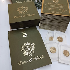 May include: Olive green wedding invitations with gold foil lettering and a floral crest. The invitations are stacked in piles, with matching wax seals on small white cards. The text "Emine & Mustafa" is visible.