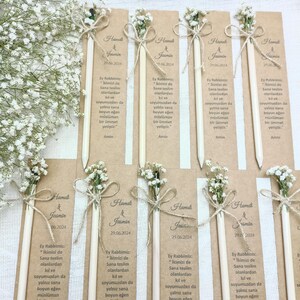 Może przedstawiać: Kolekcja spersonalizowanych oł&oacute;wk&oacute;w z kwiatowymi akcentami i kokardkami ze sznurka. Każdy oł&oacute;wek ozdobiony jest małym bukietem białych kwiat&oacute;w i etykietą z tekstem. Oł&oacute;wki ułożone są na jasnobrązowej powierzchni.