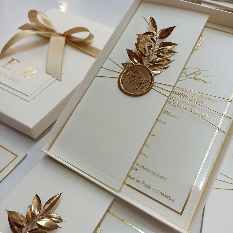 May include: Ivory wedding invitation set with gold accents. The invitation features gold foil text and a gold wax seal with the initials "EB". A gold ribbon ties the invitation box. The set includes additional cards.