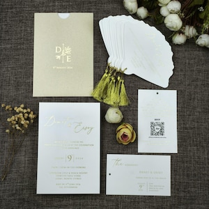 May include: Wedding stationery set with gold foil lettering on white and cream card. Includes invitation, RSVP, ceremony card, and fan programs with gold tassels. Floral accents enhance the elegant design.