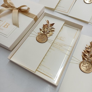 May include: Ivory wedding invitations with gold accents, presented in a white box tied with a gold ribbon. The invitation features gold foil lettering and a gold seal with initials. The box is open, revealing the invitation and a gold leaf embellishment.