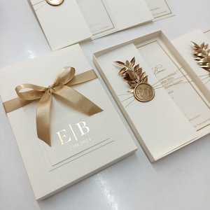 May include: Ivory wedding invitation set with gold accents. The invitation box is tied with a gold ribbon and features a clear overlay with initials and a date. Other invitations have gold leaf and seal details.