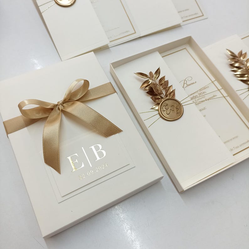 May include: Ivory wedding invitation set with gold accents. The invitation box is tied with a gold ribbon and features a clear overlay with initials and a date. Other invitations have gold leaf and seal details.
