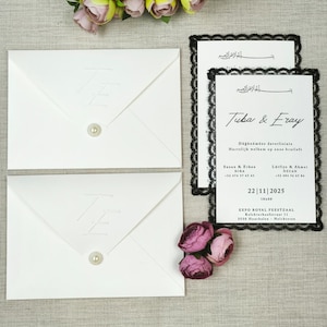 May include: Elegant white wedding invitations with black lace edging and matching envelopes. The invitations display the names "Tuba & Eray" and event details. Decorative flowers and a pearl add to the design.