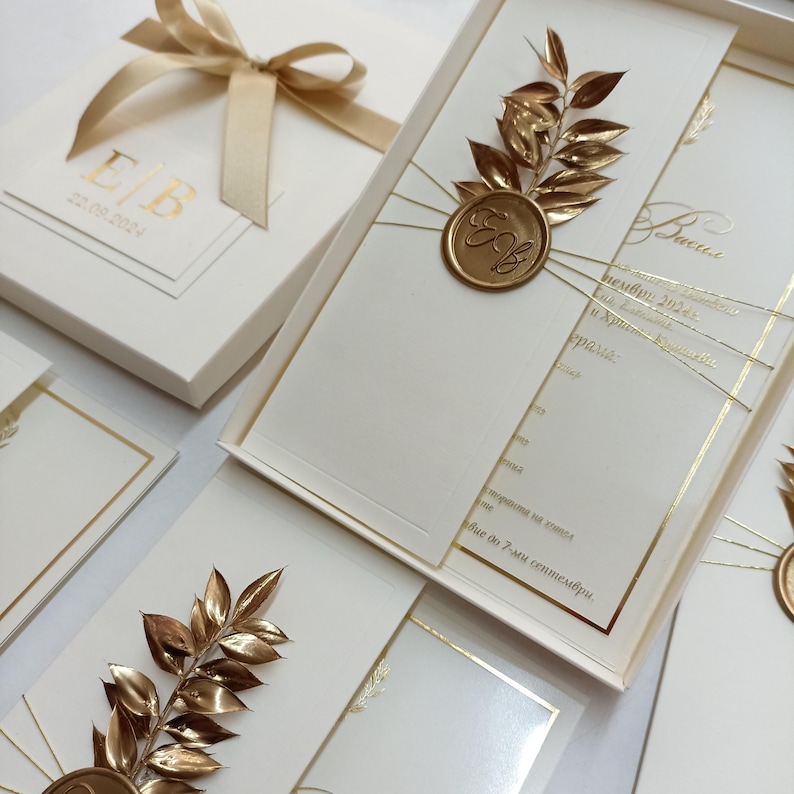 May include: Ivory wedding invitations with gold accents. Each invitation features a gold leaf design and a gold wax seal with the initials "E/B". The invitation boxes are tied with a gold ribbon. The date "22.08.2024" is printed on the box.