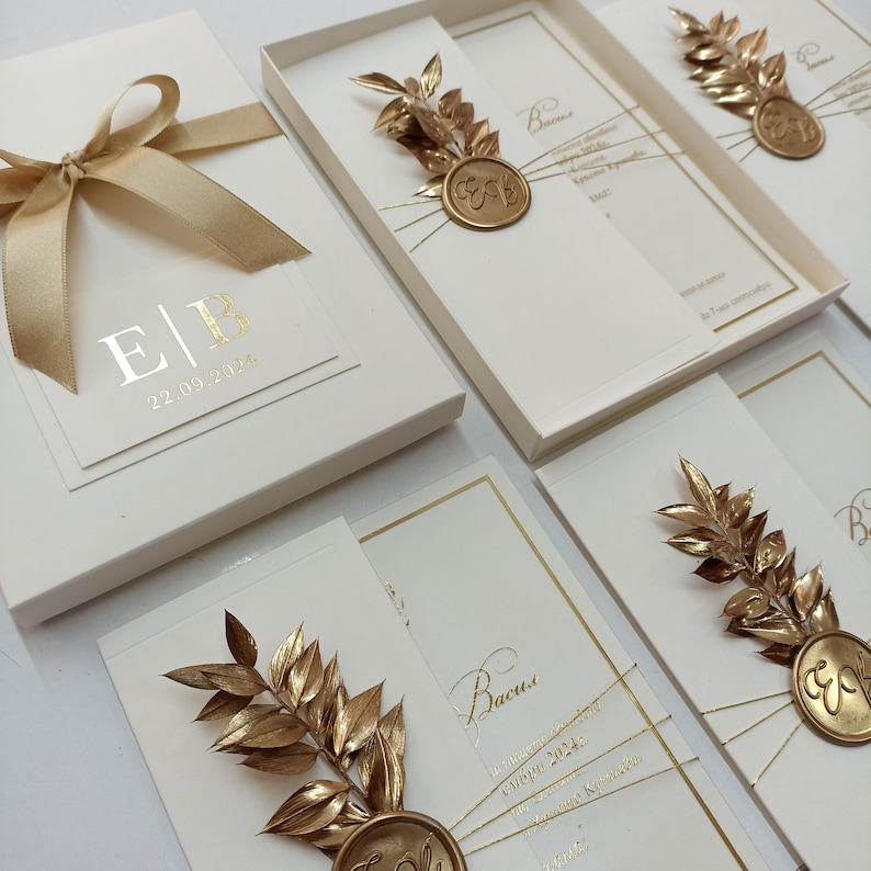 May include: Ivory wedding invitations with gold foil accents. Each invitation features a gold leaf and seal design. The invitation box has a gold ribbon and the initials "E|B" with the date "22.09.2024".