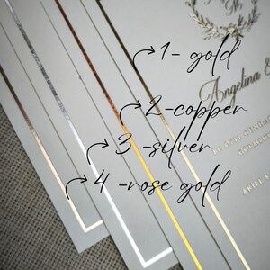 May include: A stack of invitation cards with gold, copper, silver, and rose gold foil accents. The cards have a white border and feature the names "Angelina &" and other text in gold. The image is angled.