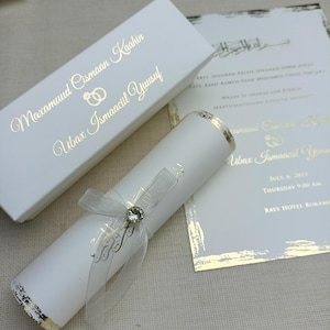 Peut inclure: Ensemble de faire-part de mariage blanc élégant. Comprend un parchemin noué avec un ruban et un bijou, une boîte assortie et une carte imprimée. Lettrage et accents dorés pour une touche de luxe. Le texte comprend les noms et les détails du mariage.
