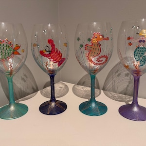 May include: Four hand-painted wine glasses with sea creature designs. Each glass features a different aquatic theme: a fish, a lobster, a seahorse, and a mermaid. The stems are painted in various colors, including teal, blue, and purple, with glitter accents.