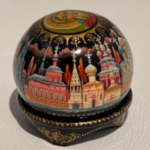 May include: A hand-painted, spherical decorative ornament with a black background. The ornament features a detailed cityscape with colorful buildings and gold accents. It sits on a matching black base with gold filigree.