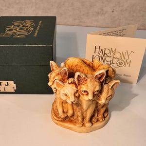 May include: A Harmony Kingdom figurine of five stylized kittens in shades of tan and orange. The figurine is next to a dark green box and a card with the Harmony Kingdom logo.