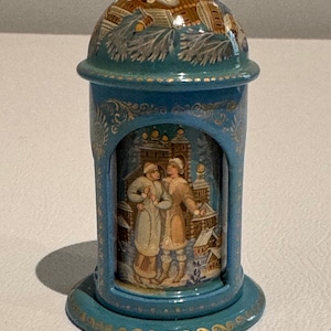 May include: A teal-colored decorative container with a domed lid. The container features a painted scene of two figures in winter clothing. Gold accents adorn the edges and base of the container, adding a touch of elegance to the design.