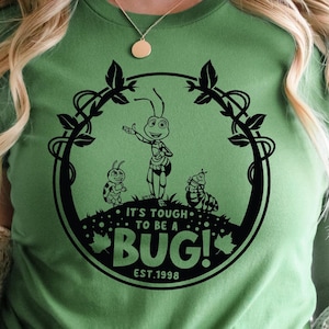 以下が含まれることがあります： 円形のフローラルデザインの中に、漫画の虫の黒いグラフィックが描かれた緑色のTシャツ。「IT'S TOUGH TO BE A BUG! EST. 1998」の文字もプリントされています。Tシャツは無地の緑色です。