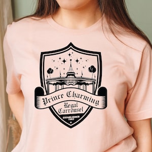 以下が含まれることがあります： 黒いグラフィックデザインが施された、薄いピンク色のTシャツ。デザインには、メリーゴーランドのイラスト、星、そして「Prince Charming Regal Carrousel」と「Established 1983」の文字が入った盾が含まれています。