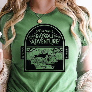 May include: A green t-shirt with a black graphic design featuring a scene from the movie "The Princess and the Frog". The graphic includes the text "Tiana's Bayou Adventure" and the years "ESTD 2024".