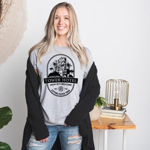 May include: A grey t-shirt with a black graphic of a hotel building with palm trees and the text "Tower Hotel, Drop In's Welcome, Established 1994".
