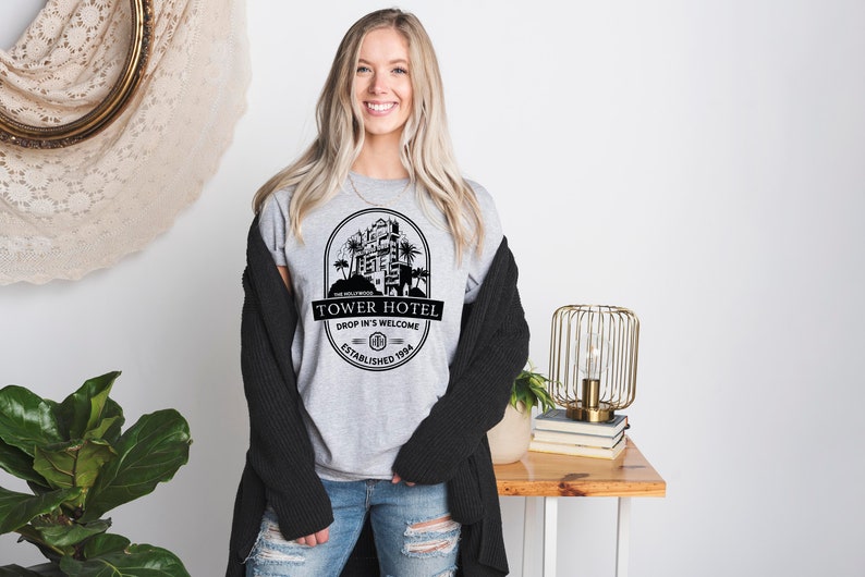 May include: A grey t-shirt with a black graphic of a hotel building with palm trees and the text "Tower Hotel, Drop In's Welcome, Established 1994".