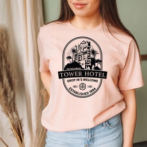 May include: A pink t-shirt featuring a black graphic of the Hollywood Tower Hotel. The graphic includes the text "The Hollywood Tower Hotel", "Drop In's Welcome", and "Established 1994".