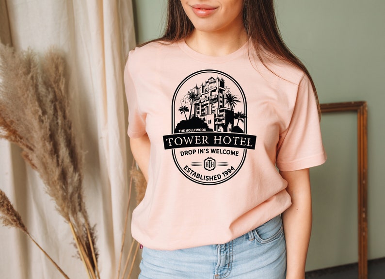 May include: A pink t-shirt featuring a black graphic of the Hollywood Tower Hotel. The graphic includes the text "The Hollywood Tower Hotel", "Drop In's Welcome", and "Established 1994".