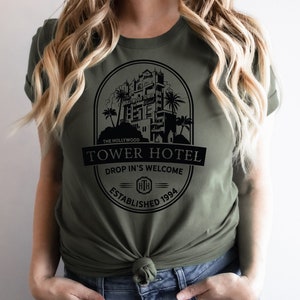 May include: A green t-shirt featuring a black graphic of the Hollywood Tower Hotel. The graphic includes the text "The Hollywood Tower Hotel", "Drop In's Welcome", and "Established 1994".